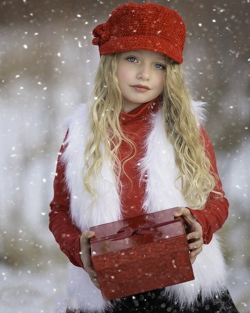 young girl with blond hair red hat white vest holding red gift standing in snow